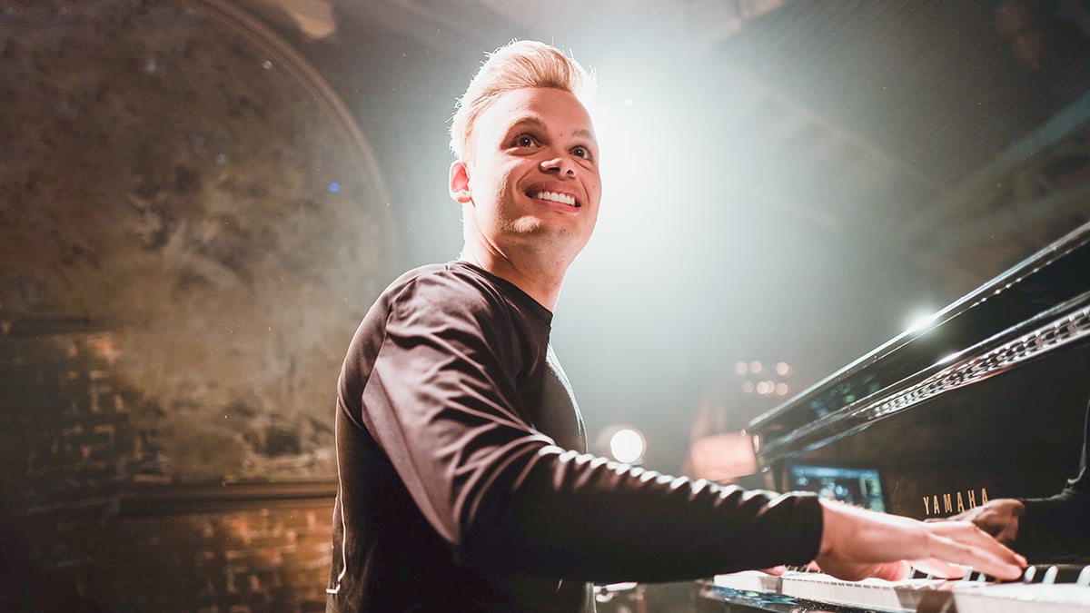 Angled-up photo of young man with blonde hair playing grand piano in well lit studio.