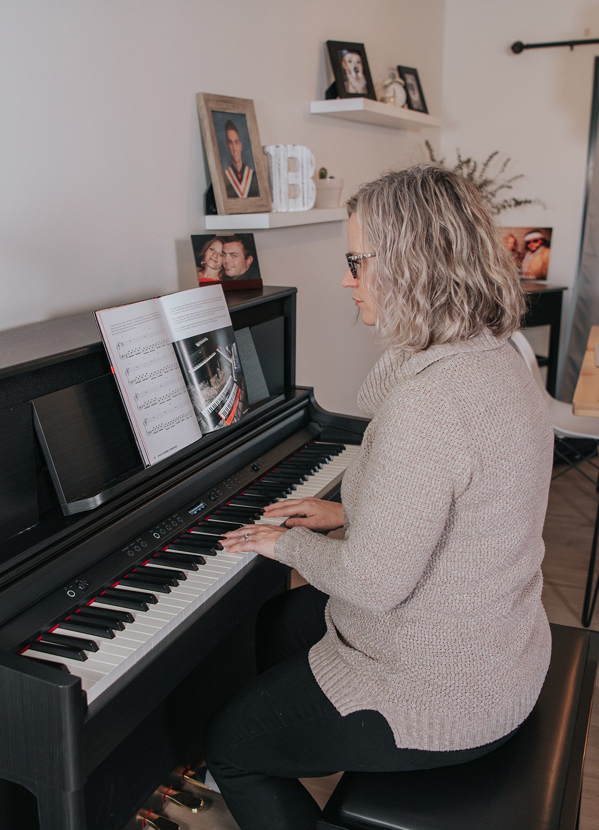 Piano practice motivation. Woman with glasses in beige sweater playing upright digital piano in home.