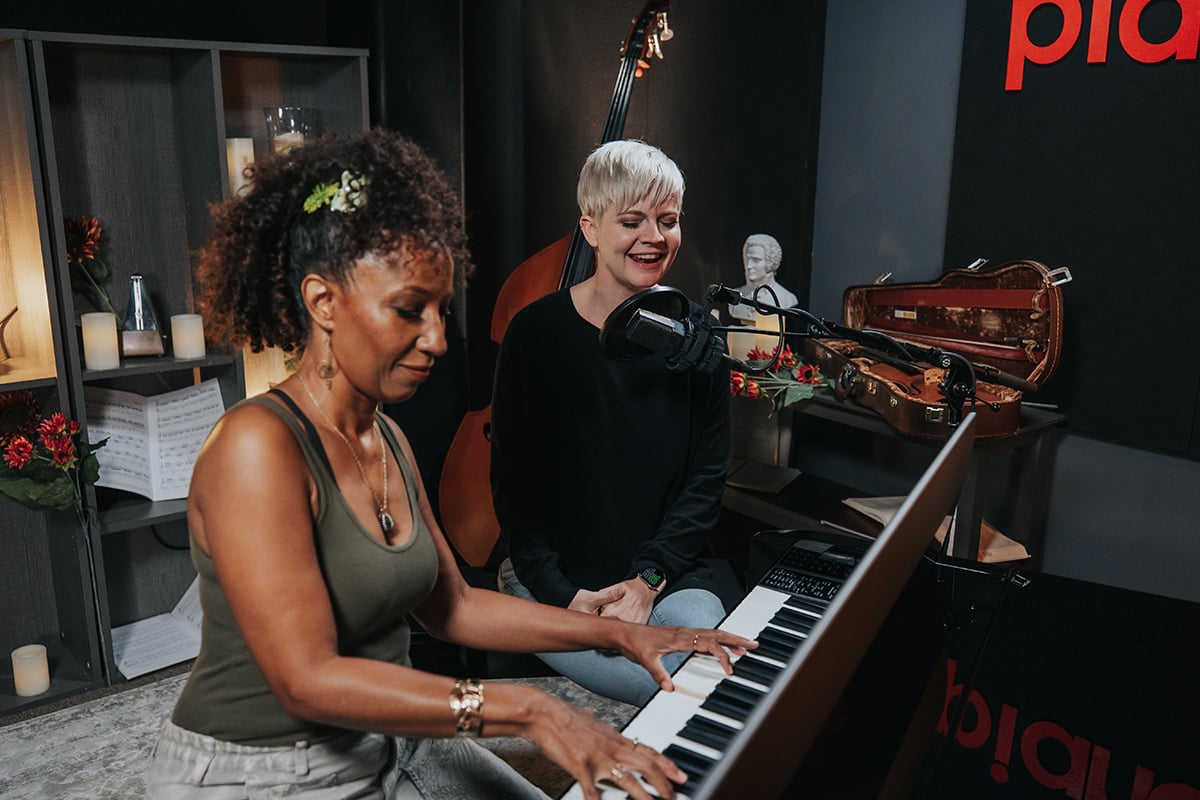 Pop piano songs. Woman with short platinum hair singing into microphone next to woman with dark curly hair playing piano.