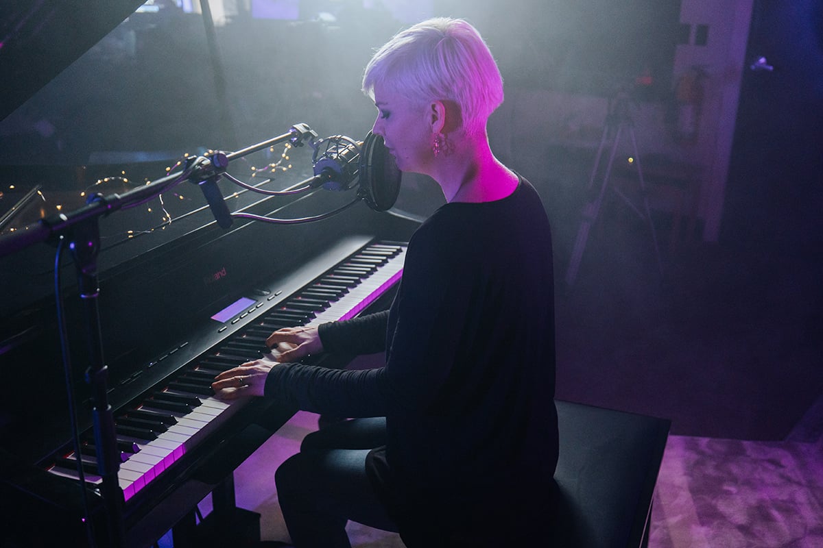 Pop piano songs. Woman with short platinum hair singing into mic and playing piano in purple lit lounge like room with fair lights and vapor.